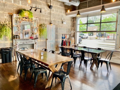 A bright, modern dining area with wooden tables, black chairs, and greenery against a brick wall. Large windows let in natural light.