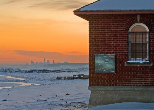 A brick building in the foreground with a snowy landscape and a city skyline at sunset in the background.
