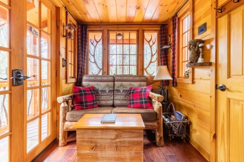 Cozy wooden cabin interior with a sofa, plaid cushions, a coffee table, and large windows letting in natural light.