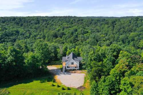 Aerial view of a modern house surrounded by lush green trees and a gravel driveway in a wooded area.