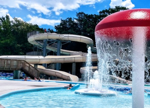 A water park scene featuring slides, a splash pad, and people enjoying the pool under a bright blue sky.