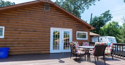 A wooden cabin exterior with a deck, table, and colorful chairs, surrounded by trees and a clear blue sky.