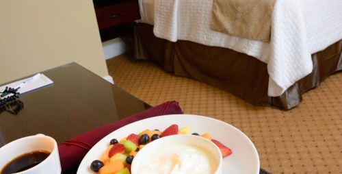 A plate of fresh fruit and yogurt on a table, with a cup of coffee and a hotel bed in the background.