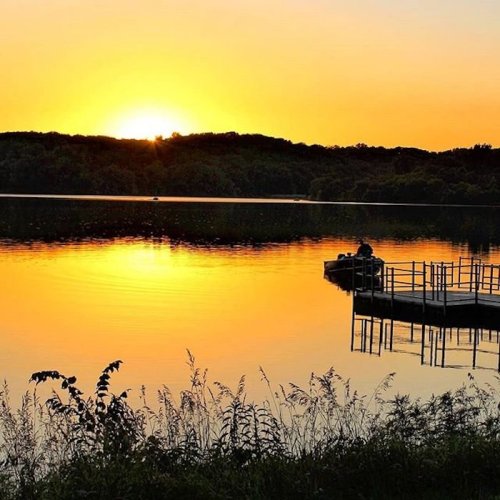 A serene sunset over a calm lake, with a dock and lush greenery in the foreground.