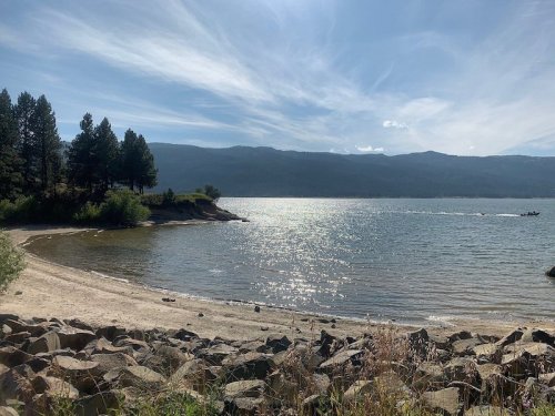 A serene lakeside view with sandy shore, rocky foreground, and mountains under a blue sky with wispy clouds.