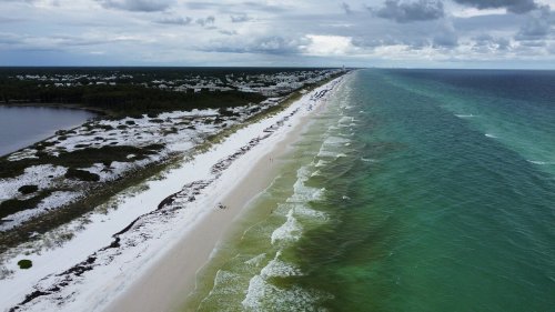 Aerial view of a sandy beach with turquoise waters, surrounded by greenery and a distant coastline under cloudy skies.
