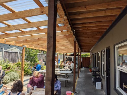 Outdoor seating area with wooden beams, tables, and people enjoying their time in a sunny, garden-like setting.