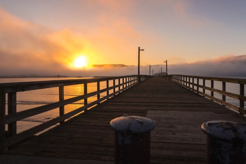 A wooden pier extends into a calm waterway, with a vibrant sunset partially obscured by fog in the background.