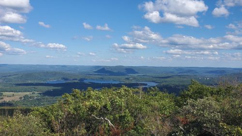 A panoramic view of rolling hills and lakes under a blue sky with fluffy clouds. Lush greenery in the foreground.