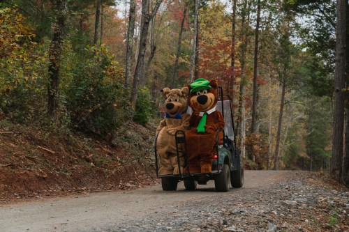 Two people in bear costumes sit in a vehicle on a gravel road surrounded by colorful autumn trees.