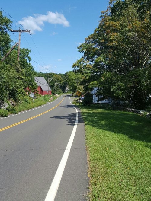 A winding road lined with trees and a grassy area, leading towards a distant red barn under a clear blue sky.