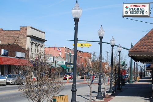 A quaint downtown street with lamp posts, shops, and bare trees under a clear blue sky. Sign for Bedford Floral Shoppe visible.
