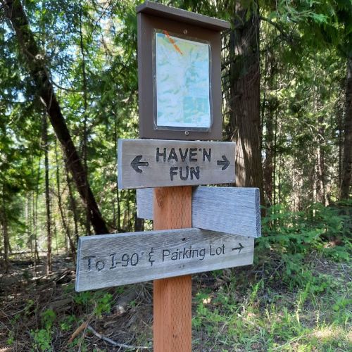 Wooden signpost in a forest with directions to "HAVE'N FUN" and "To I-90 Parking Lot," featuring a map above.