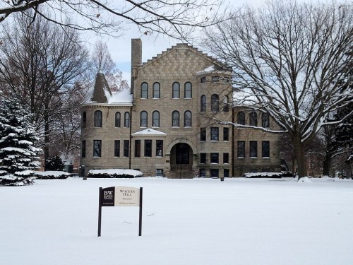 Historic stone building, Whittier Hall, surrounded by snow-covered trees and a sign in front. Winter scene.