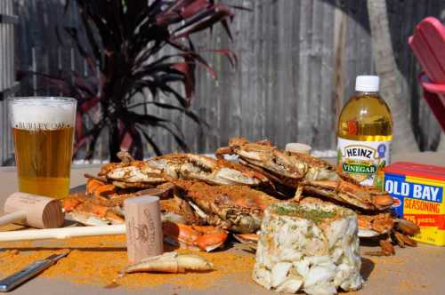 A table spread with seasoned crabs, a bottle of vinegar, Old Bay seasoning, a beer, and crab mallets.