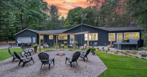 A cozy black house surrounded by trees, featuring a fire pit and seating area in a landscaped yard at sunset.