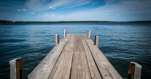 A wooden pier stretches toward the horizon on Okoboji Lake, Iowa. The water is calm. The sky overhead is blue.
