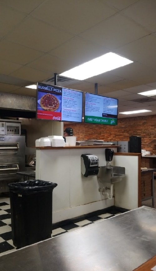 Interior of a pizza restaurant featuring menu boards, a sink, and a trash can, with a checkered floor.