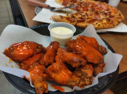 A plate of spicy chicken wings with a side of ranch dressing, with a pizza in the background.