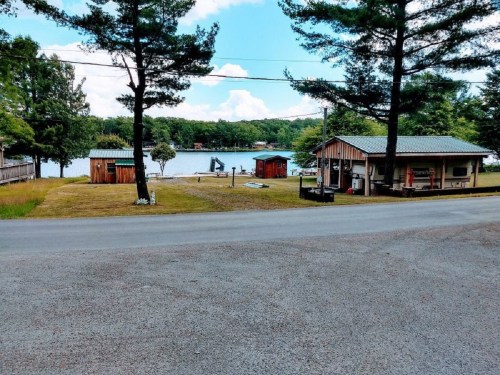A scenic view of a lake with cabins and trees, set against a clear blue sky. A road runs in the foreground.