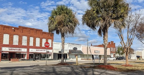 A small town street with palm trees, storefronts, and a stop sign under a partly cloudy sky.
