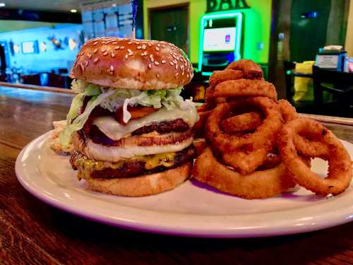 A sesame seed burger stacked with lettuce, tomato, cheese, and a side of crispy onion rings on a plate.