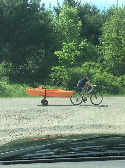A person rides a bicycle pulling an orange kayak on a wheeled trailer through a grassy area.