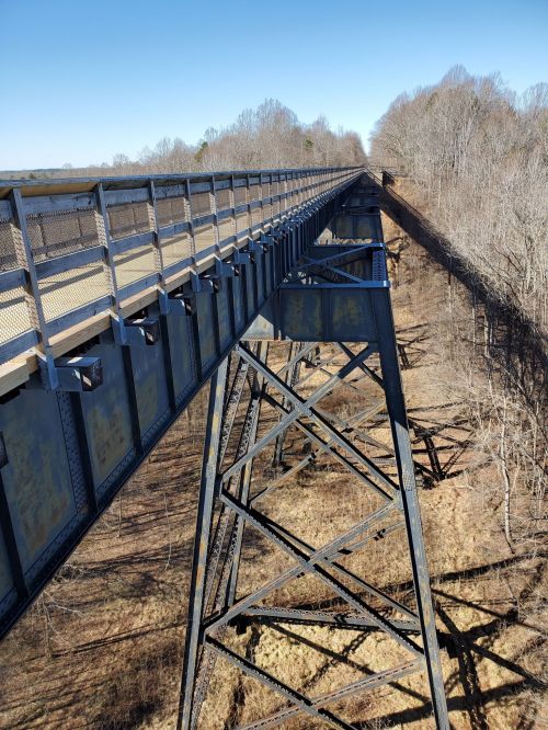 A high metal bridge spans a wooded area, with clear blue skies and trees lining the sides.