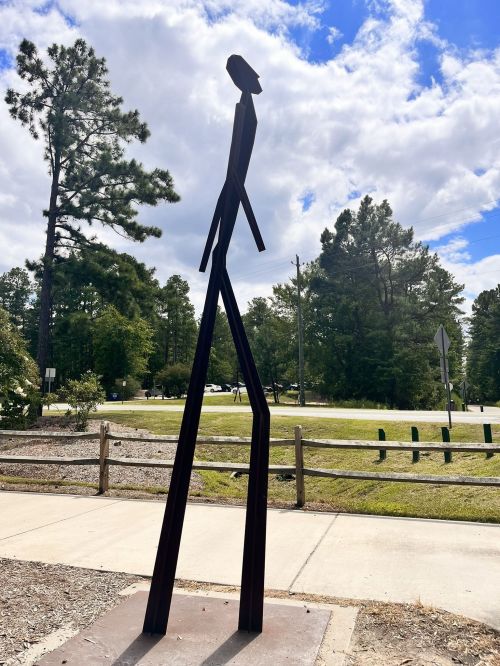 A tall, abstract metal sculpture of a person walking, set against a backdrop of trees and a blue sky.