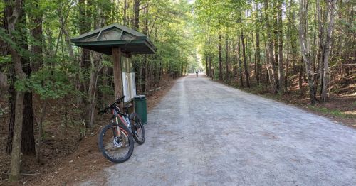 A gravel path through a wooded area, with a bike parked at a shelter and a person walking in the distance.