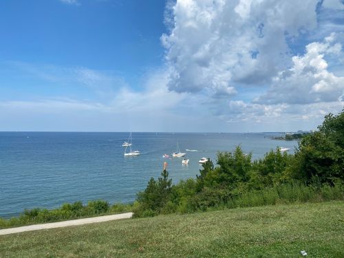 A serene view of a blue lake with sailboats, under a partly cloudy sky and surrounded by green grass and trees.
