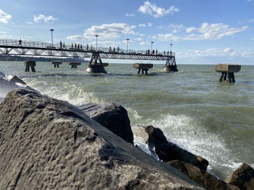 A pier extends over choppy waters, with people walking along it under a blue sky with scattered clouds.