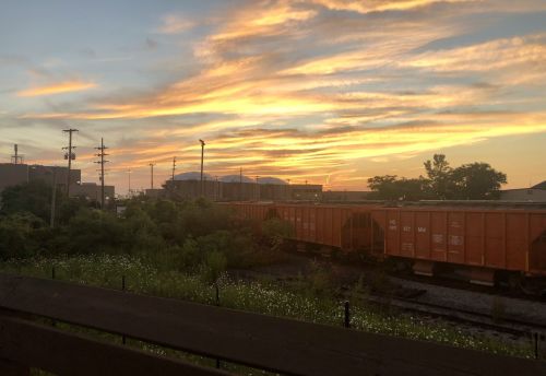 A vibrant sunset over a train yard, with orange clouds and silhouettes of buildings and trees in the background.