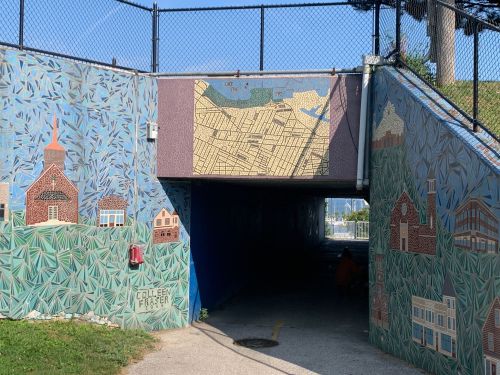Colorful mural in a tunnel featuring buildings and a map, with a blue wall and a fence above.