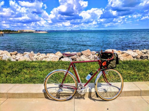 A red bicycle with a bag parked by a rocky shoreline, overlooking a blue lake and a skyline in the distance.