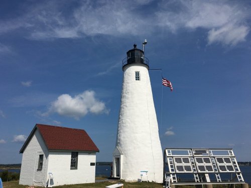A white lighthouse with a red roof, solar panels, and an American flag, set against a blue sky and water.