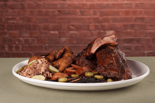 A platter of smoked meats including ribs, pulled pork, and sausage, garnished with pickles and served on a green tablecloth.