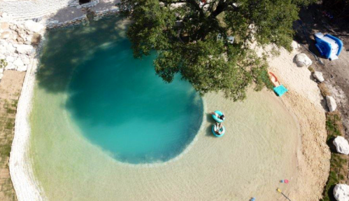 Aerial view of a clear blue swimming hole surrounded by sandy areas and a large tree. Two people float on inflatable rings.