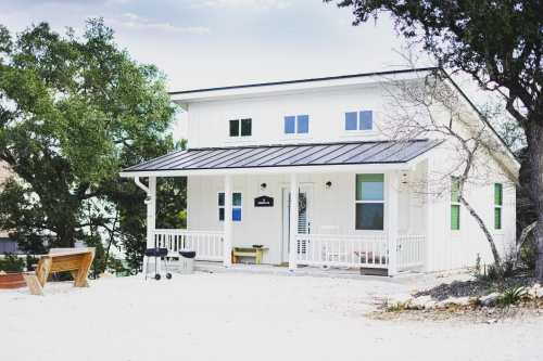 A modern white house with a metal roof, surrounded by trees and a gravel path, featuring a porch and outdoor seating.