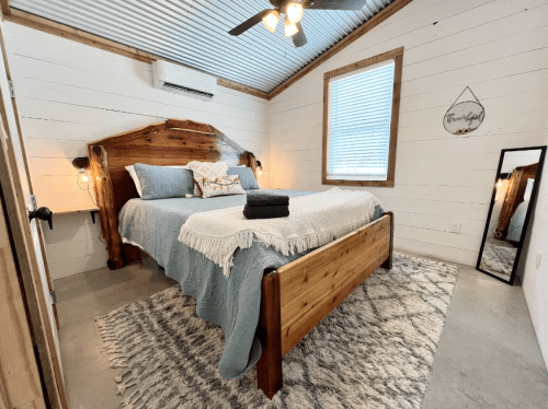 Cozy bedroom with a wooden bed, blue bedding, a mirror, and a textured rug against a white shiplap wall.