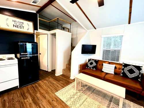 Cozy interior of a small home with a sofa, kitchen area, and stairs leading to a loft, featuring a welcoming sign.