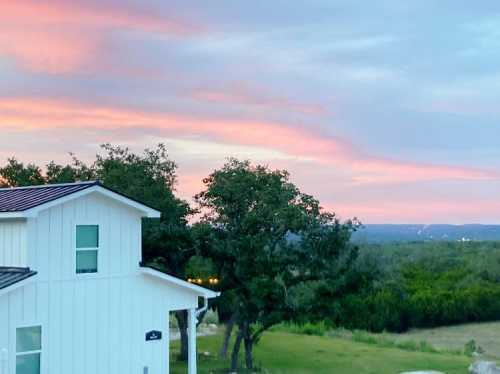 A white house with a sloped roof surrounded by trees, set against a colorful sunset sky.