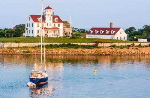 A sailboat floats in calm water near a white lighthouse and a coastal house with red roofs.