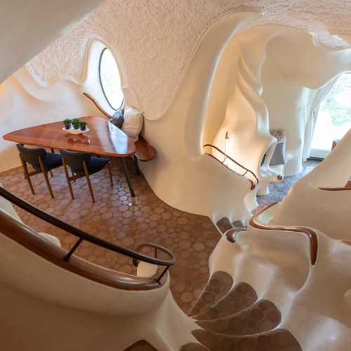 Interior of a uniquely shaped room with smooth, organic walls, a wooden table, and a round window.