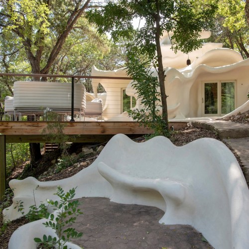 A unique, organic-shaped white house surrounded by trees, with a curved pathway and outdoor seating area.