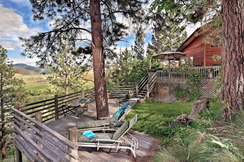 A cozy wooden cabin surrounded by trees, featuring a deck with lounge chairs and a scenic view of rolling hills.