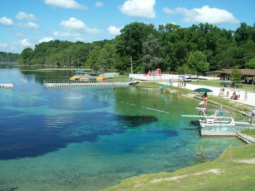 A scenic lake view with clear water, a sandy beach, and people enjoying recreational activities under a blue sky.