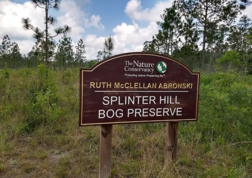 Sign for Ruth McClellan Abronski Splinter Hill Bog Preserve, surrounded by greenery and trees under a cloudy sky.