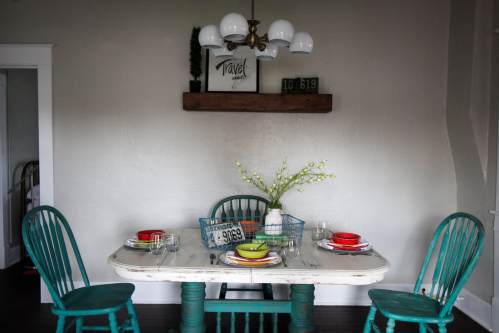 A cozy dining area with a rustic table, teal chairs, and a decorative shelf featuring plants and travel-themed decor.