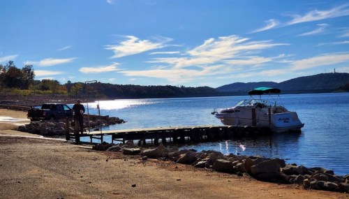 A serene lakeside scene with a boat dock, a parked truck, and a person walking along the shore under a clear blue sky.